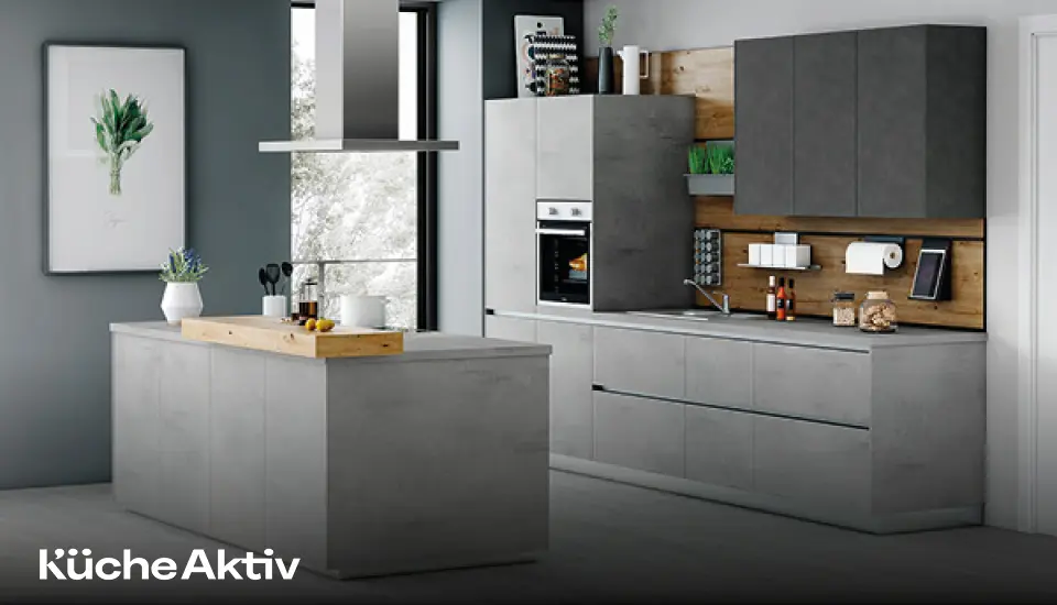 Moderne Küche in grauer Betonoptik mit Insel, Hängeschränken und Holzrückwand, ausgestattet mit Backofen, Dunstabzugshaube und dekorativen Accessoires vor großem Fenster